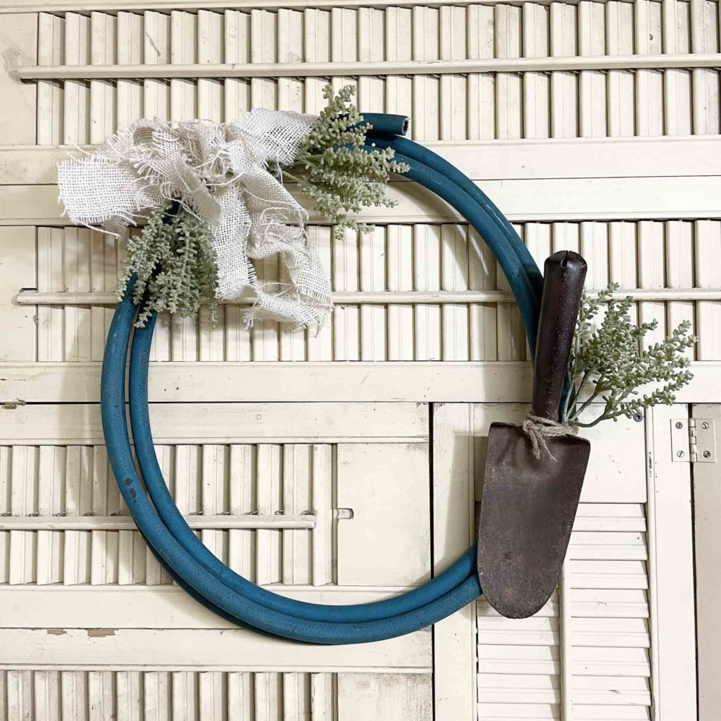 Teal garden hose looped in a wreath form with greenery, burlap, and garden shovel.