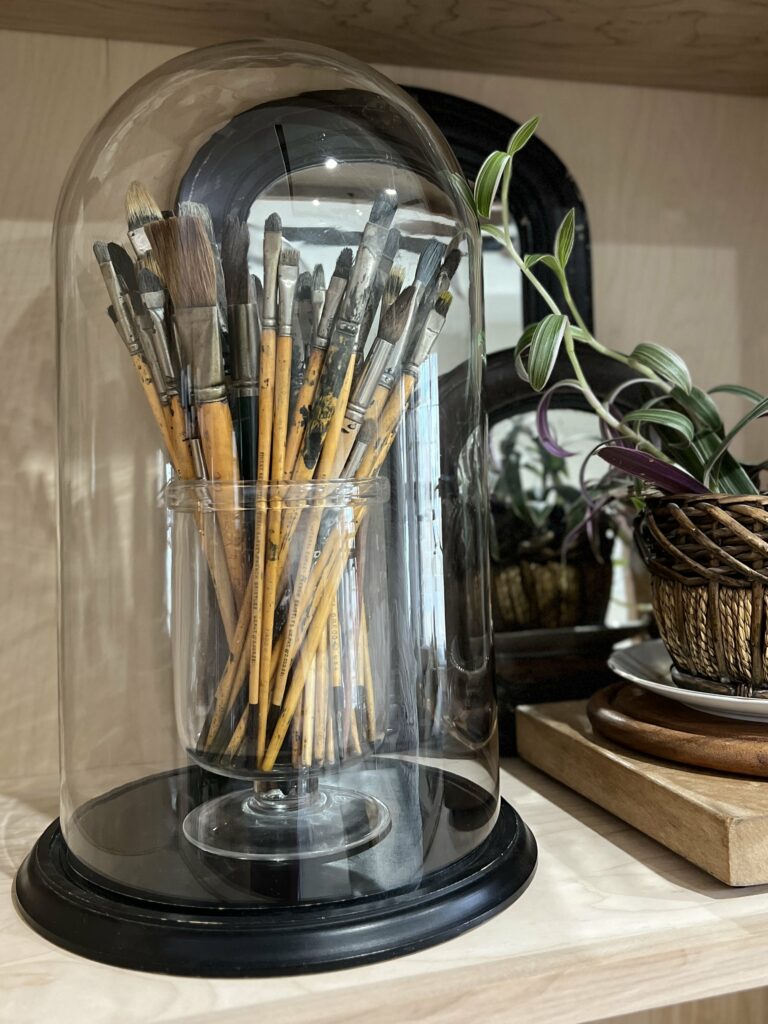 Collection of paint brushes in a vase under a huge cloche.