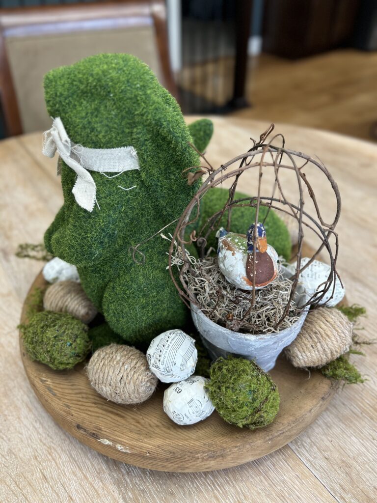 Topiary with a paper mache candy duck inside. Topiary pot is sitting amongst repurposed easter eggs and moss bunies