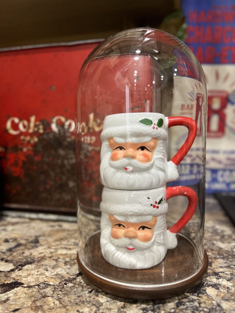 2 santa mugs stacked underneath a cloche with a cola cooler in the background
