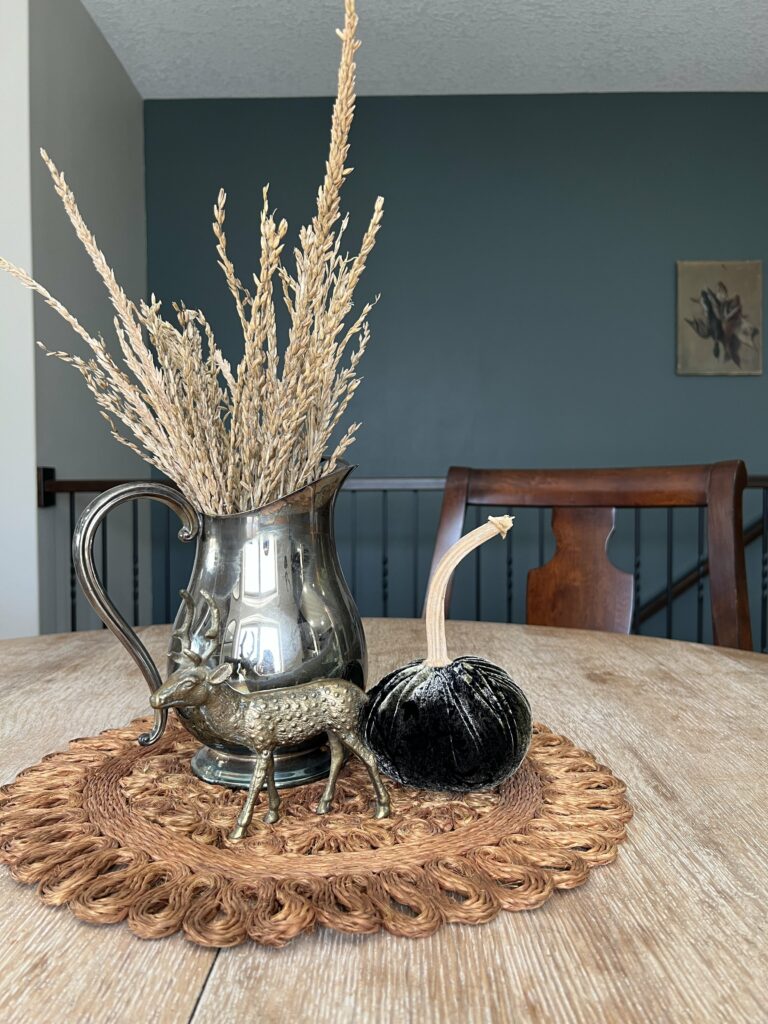 a round placemat topped with a silver pitcher, brass deer and pumpkin