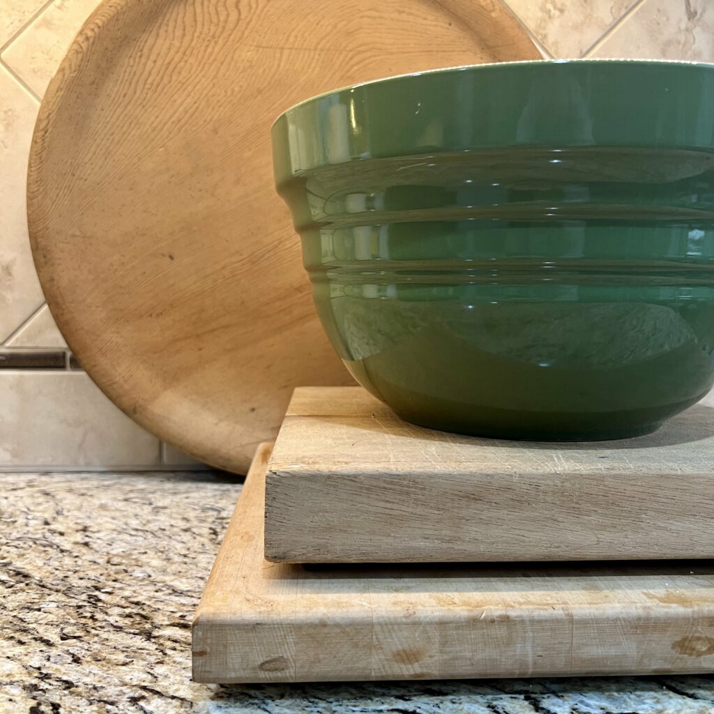 Layers - Square cutting boards stacked with a green bowl on top and a round bread board in the back.
